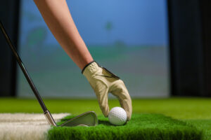 Golfer Hand with Glove Placing Ball on Artificial Turf in Indoor Golf Simulator Training Facility, golf shop Pekin IL, golf store near Pekin IL, golf clubs Pekin IL, golf equipment Pekin IL, golf pro shop Pekin IL, custom club fitting Pekin IL, golf simulator Pekin IL, golf lessons Pekin IL, used golf clubs Pekin IL, golf accessories Pekin IL, golf club fitting near Pekin IL, indoor golf Pekin IL, golf club repair Pekin IL, buy golf clubs Pekin IL, golf retail store Pekin IL, golf balls Pekin IL, golf apparel Pekin IL, boutique golf shop Illinois, local golf store near Pekin, golf store Central Illinois