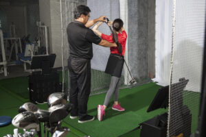 A woman practicing at a golf studio and a man with a coach, golf shop Pekin IL, golf store near Pekin IL, golf clubs Pekin IL, golf equipment Pekin IL, golf pro shop Pekin IL, custom club fitting Pekin IL, golf simulator Pekin IL, golf lessons Pekin IL, used golf clubs Pekin IL, golf accessories Pekin IL, golf club fitting near Pekin IL, indoor golf Pekin IL, golf club repair Pekin IL, buy golf clubs Pekin IL, golf retail store Pekin IL, golf balls Pekin IL, golf apparel Pekin IL, boutique golf shop Illinois, local golf store near Pekin, golf store Central Illinois